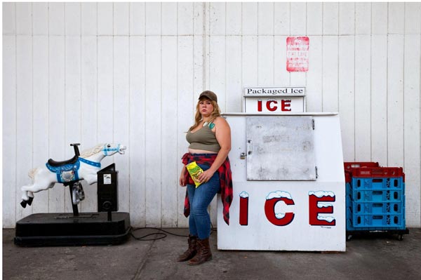 A woman in a cap and jeans stands next to an ICE freezer and a blue and white horse ride, evoking a Genevieve Gaignard tableau. She holds a snack bag, wearing a green top and plaid shirt tied around her waist, set against a white wall.