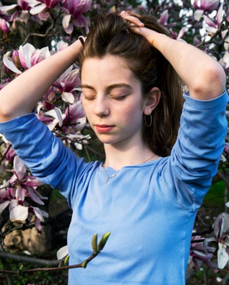 A person in a blue shirt, evoking family memories, stands with closed eyes and hands behind head amidst the blooming pink and white magnolia flowers.