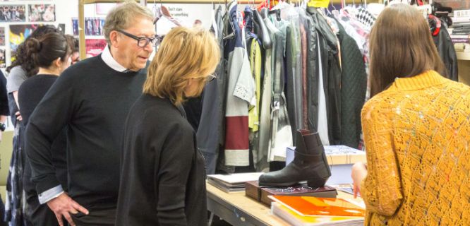 Two people, a man and a woman, examine a black boot on a table covered with books and folders, perhaps delving into insights from their Footwear Course at MassArt. In the background, racks of colorful clothing can be seen. Nearby, someone in a vibrant orange sweater is partially visible.