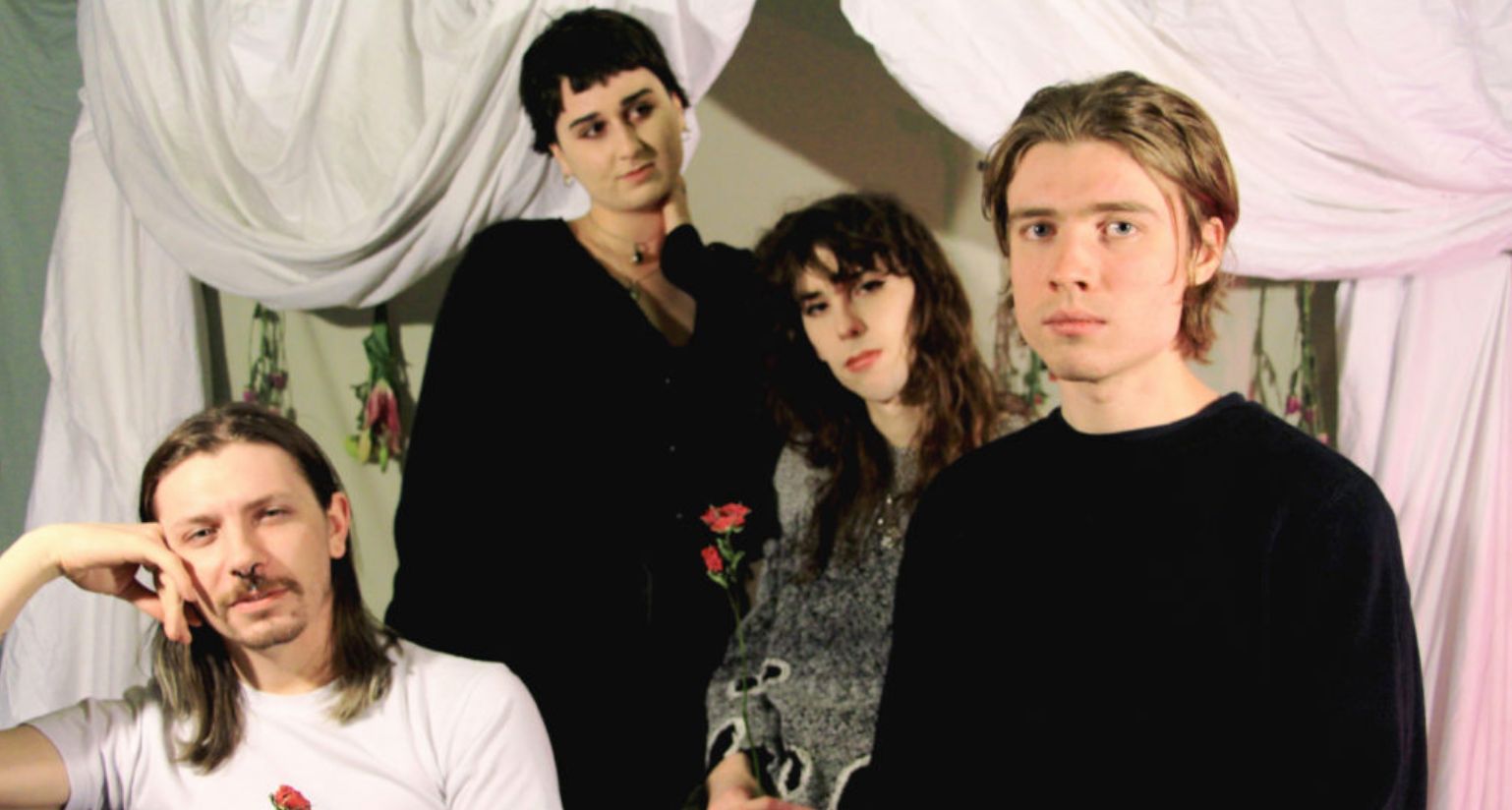 Four people, members of an experimental metal band, pose in front of draped white fabric. Three stand while one sits, resting his head on his hand. Their eclectic outfits include a light floral sweater and a black one. The atmosphere is relaxed and artistic, celebrating queer identities.