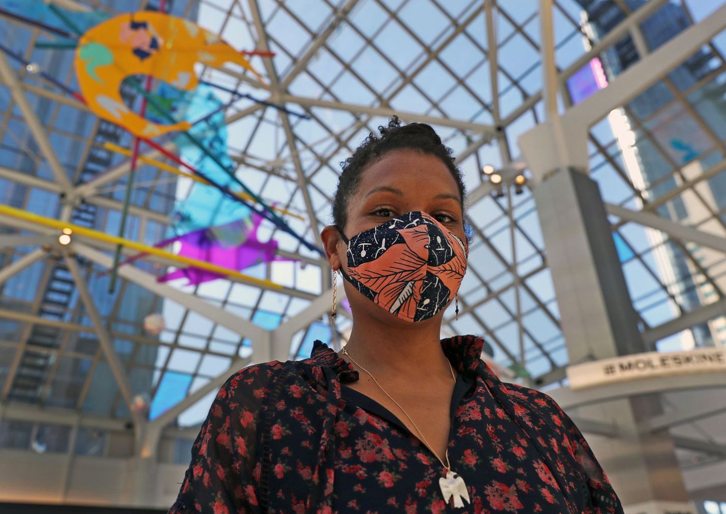 A person wearing a floral mask and floral-patterned blouse stands inside a modern glass-ceiling building, part of the intriguing Pru Installation. Colorful art pieces hang overhead. With short hair, earrings, and a necklace, this artist invites onlookers to look up in awe.