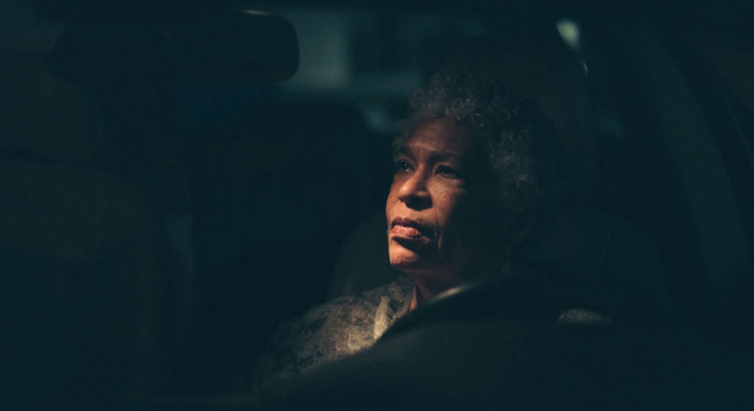 An elderly person with curly hair gazes contemplatively out of a car window at night, dimly lit from the front. Inspired by Amani Willetts evocative visuals, they seem to be capturing the essence of a Great American Road Trip under a starlit sky.