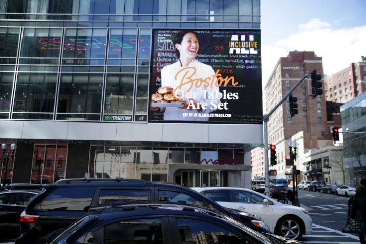 A busy city street with cars in front of a modern building. A large digital billboard showcases a smiling person and text for an all-inclusive Our Tables Are Set Boston dining campaign. The urban scene is framed by high-rise buildings, emphasizing vibrant Boston tourism.