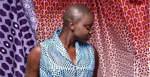 A person with a shaved head wears a sleeveless, blue patterned top, standing against a colorful, geometric-patterned fabric background in shades of purple, red, and white. They are looking to the side, embodying an artful blend of creativity and sustainability.