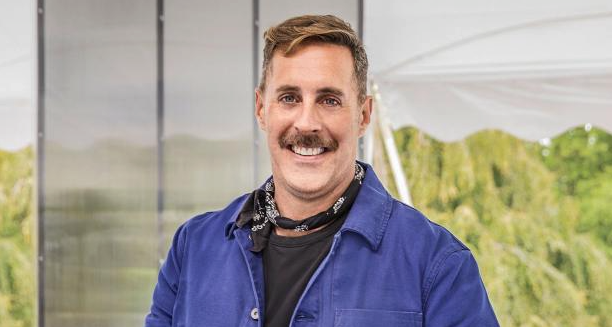 A person with a mustache smiles at the camera, exuding confidence like one of the contestants, wearing a blue jacket over a black shirt with a bandana around the neck, standing outdoors with greenery and a white tent in the background.