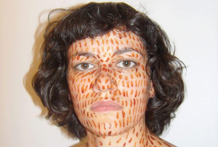 A person with short, curly dark hair, reminiscent of Zehra Khans multidisciplinary style, has their face adorned with spontaneous reddish-orange vertical brushstroke patterns. They stand against a plain white background, gazing directly at the camera with a neutral expression.