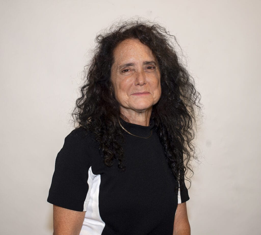 A person with long, curly hair wearing a black and white shirt stands in front of a plain background, smiling gently, embodying the spirit of fine arts.