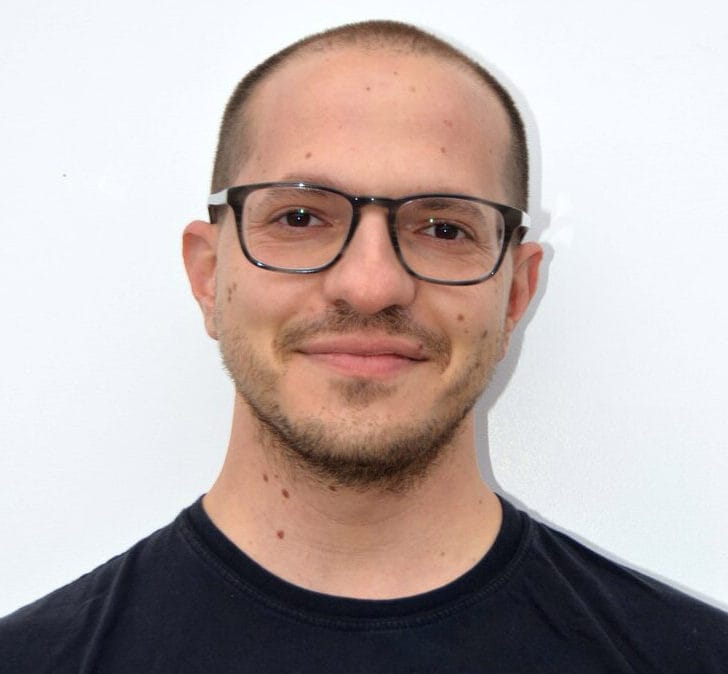 A person with short hair and glasses is smiling slightly at the camera. They are wearing a black t-shirt that seems to aid in projecting a sleek look, standing against a plain white background.