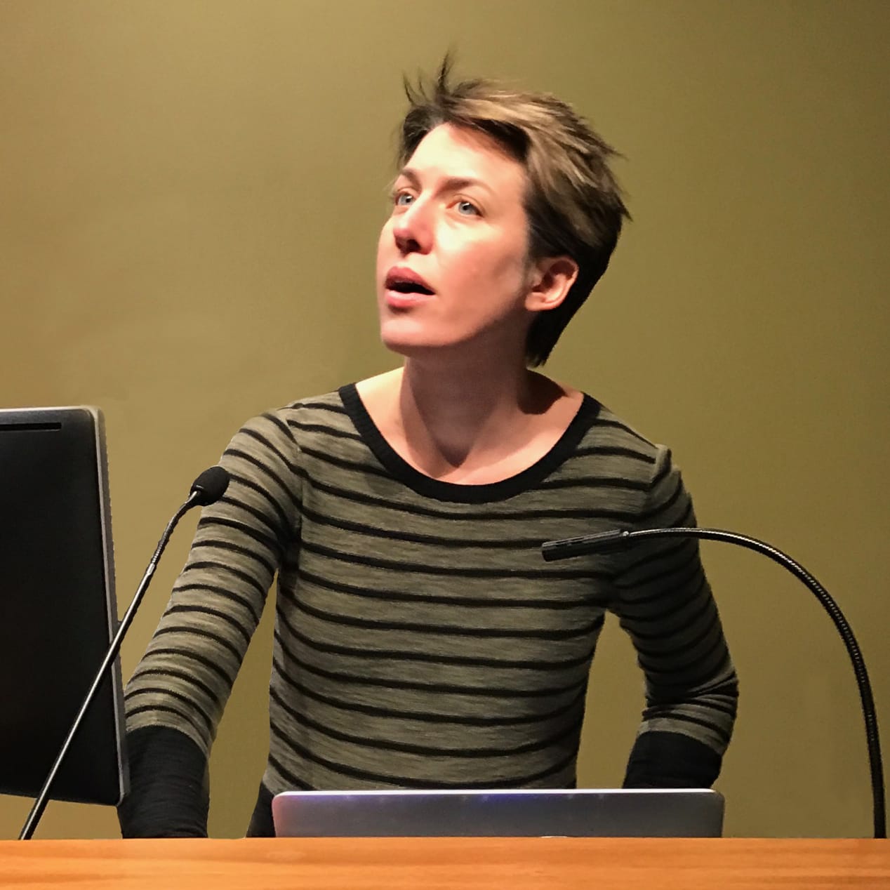 A person with short hair, in a striped long-sleeve shirt, stands at a podium with two microphones and a laptop, looking upward. The background is a plain beige wall. The scene appears to be during a presentation or lecture.