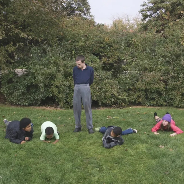 On a grassy lawn, a person stands with hands behind their back as four colorful graduates playfully crawl on the grass. Bushes and trees provide a lush, natural backdrop.