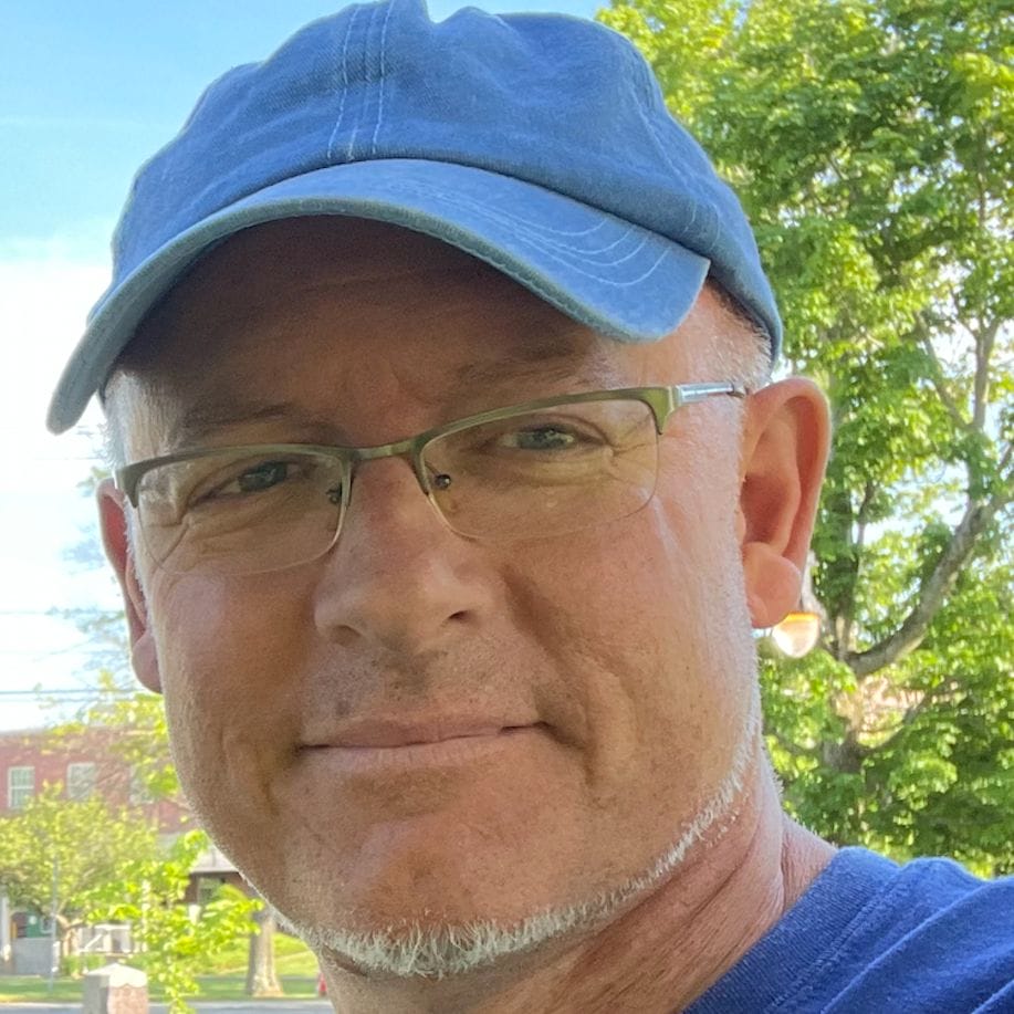 A man wearing a blue cap and glasses is smiling outdoors, as if marking the joyful start of a new academic calendar. He has a trimmed beard and is set against a background of green trees and a bright blue sky.
