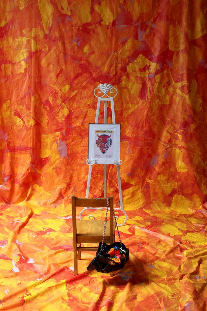 A wooden chair and a purse sit before a white easel showcasing framed art. Against the backdrop of vibrant orange and yellow abstract fabric, this captivating scene unfolds in Devils Corner, exuding creativity and charm.
