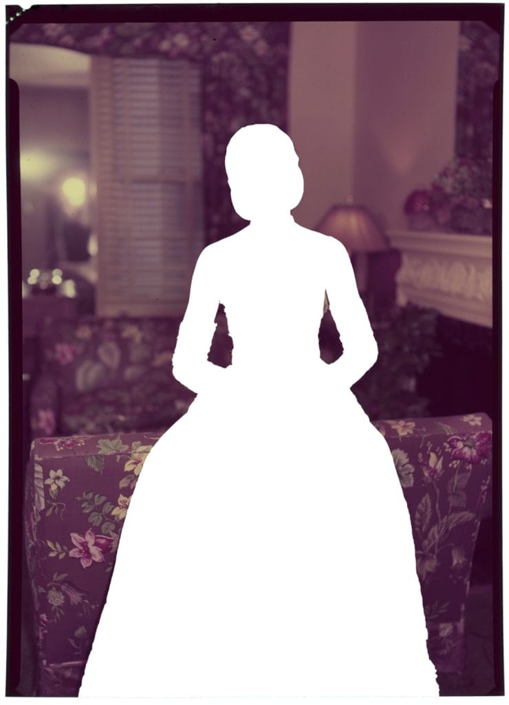 Silhouette of a person in a formal gown, poised gracefully before a floral-patterned couch. The room, softly illuminated, hints at the elegance of an Untitled series with a lamp and ornate decorations visible in the background.