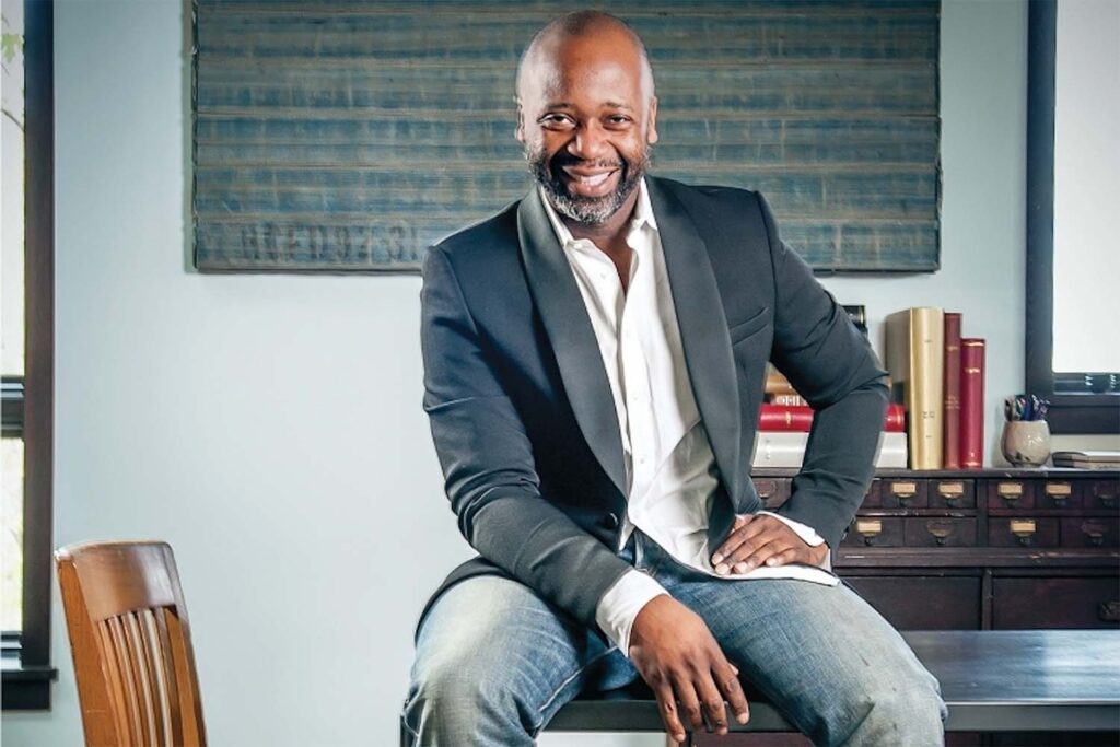 Smiling man in a dark blazer, white shirt, and jeans sits on a desk in a modern office with shelves and books in the background, embodying the confidence of a 2021 commencement speaker.