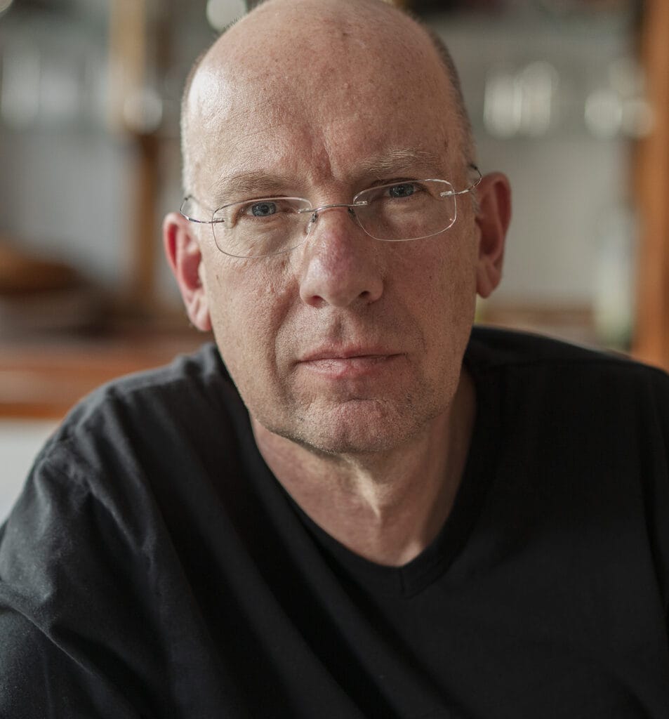 Meet the Team: A middle-aged man wearing glasses and a black shirt looks directly at the camera with a neutral expression. The background is blurred, capturing his composed presence as part of our dedicated Admissions Office.