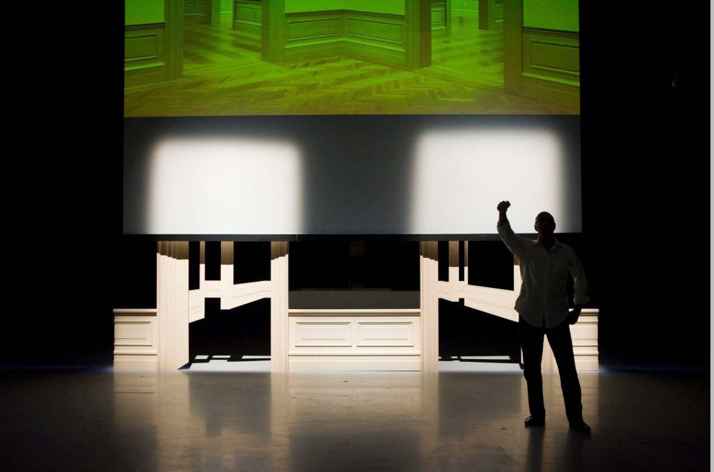 A person stands in silhouette on a dimly lit stage, raising one hand. Behind them, an abstract architectural image with green tones glows on a large screen—an ode to Fictions Truth. The stage is adorned with minimalist wooden structures, echoing the visionary spirit of Walid Raad.