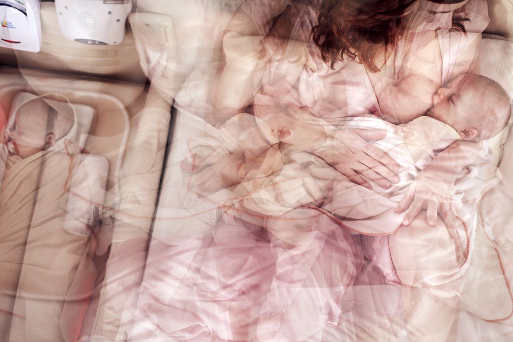 A blurry, overlapping image portrays a woman from the Designing Motherhood exhibit at MassArt Art Museum holding and breastfeeding a baby. Nearby, another baby sleeps in a seat. The scene captures the nurturing essence of caregiving and tenderness.
