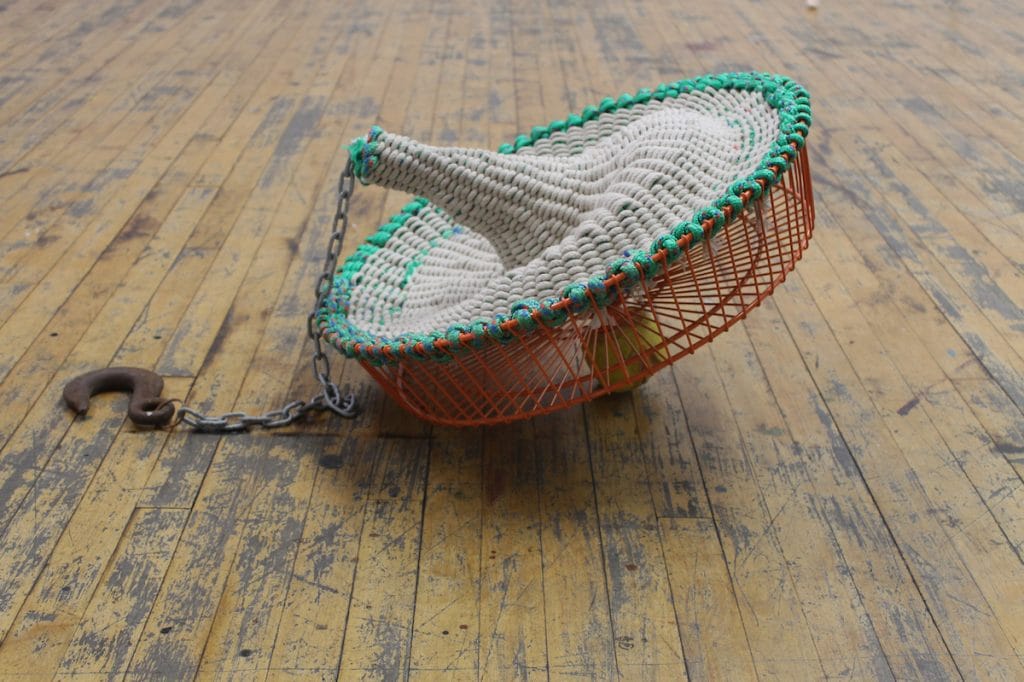 A cone-shaped sculpture of woven white material, encircled by a green-bordered orange wire frame, leans on a wooden floor like a silent ode to Yankee Pride. It is anchored by a chain with a metal hook, bridging art and legacy.