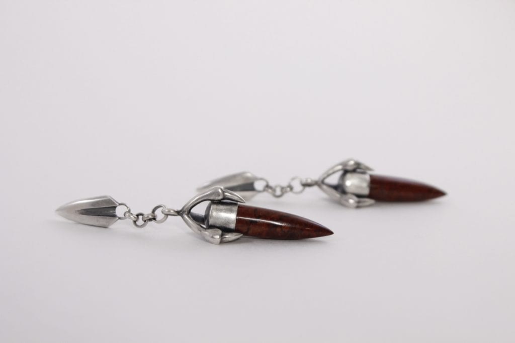 Two silver earrings with pointed red-brown stone pendants on a white background. The earrings have a metal claw-like design holding each stone, linked by small chains to a triangular metal top.