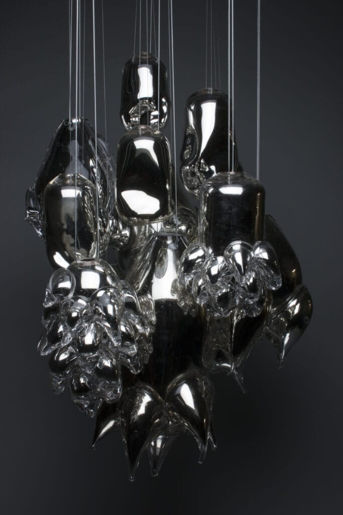 A cluster of shiny, metallic, abstract sculptures titled Silvered Distortion is suspended by thin strings. The shapes resemble organic forms with smooth, reflective surfaces and varying textures. The dark background emphasizes the sculptures glossy appearance, creating a mesmerizing visual effect.