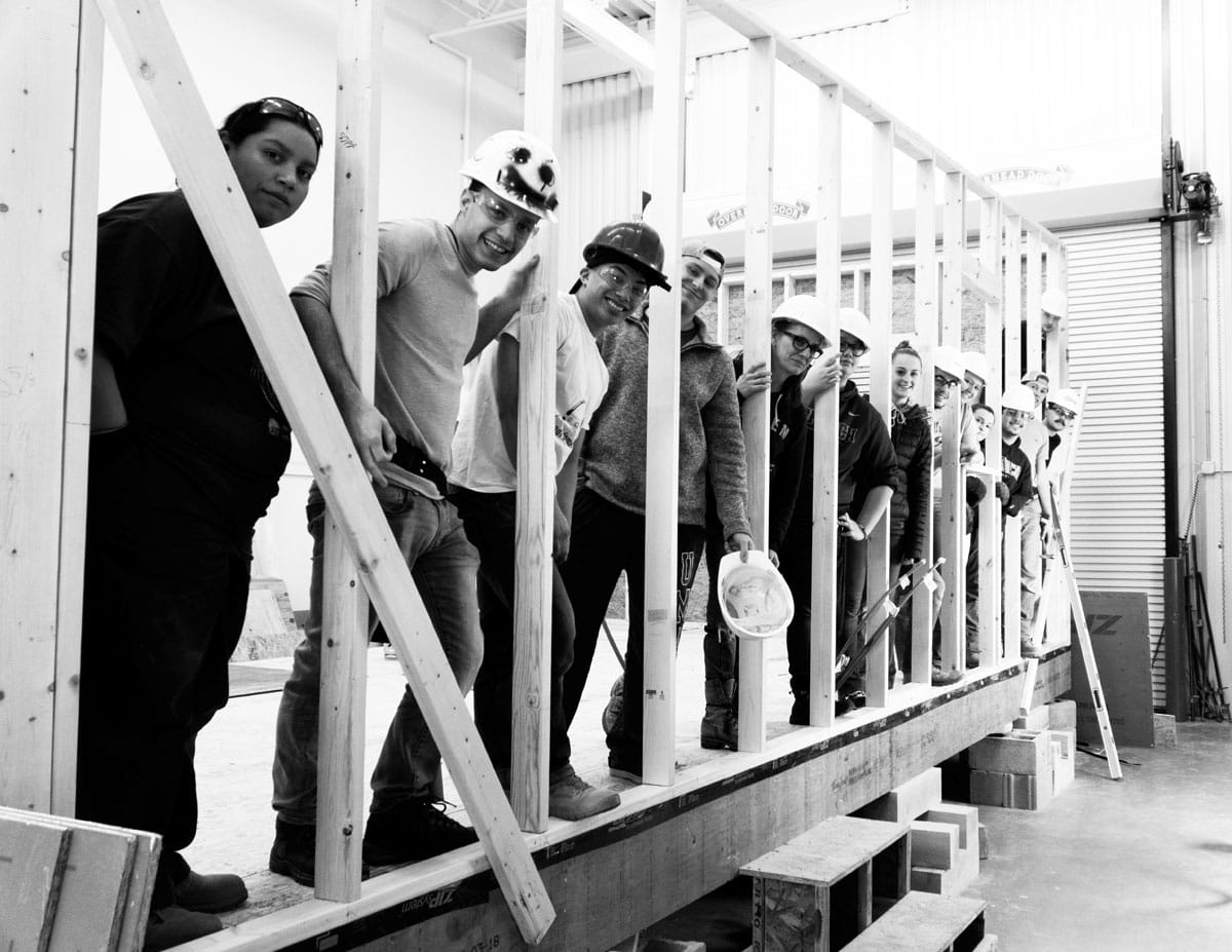 A group of construction workers stands on a wooden framework in Norwich, all wearing hard hats and holding tools. They line up, smiling and posing confidently for the camera, embodying the spirit of building Affordable Shipping Container Homes. Black and white photo.