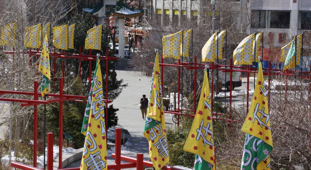 Brightly colored flags with Asian characters stand in rows on red poles, fluttering in the wind. In the background, two people walk along a path lined with trees towards a decorative green-roofed structure—a centerpiece of the Greenway Art Installation. Buildings surround the scene.