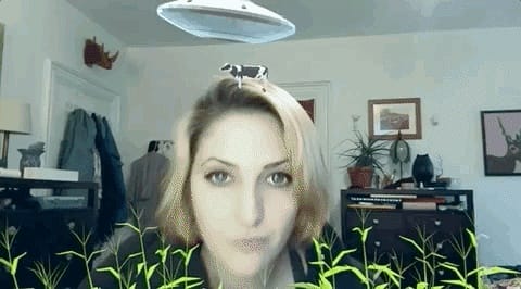 A woman with short blonde hair stands in a room, created by Snapchat Lens Creator Audrey Spencer, where a virtual UFO hovers above her head. A small cow perches on the UFO amidst green virtual grass. The scene features a plant, coat rack, and framed pictures in the background.