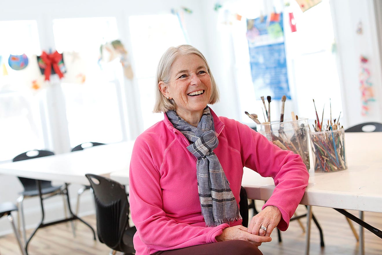 A smiling older woman with gray hair, presumably Heidi Hurley, sits in a bright classroom, wearing a pink sweater and gray scarf. The art supplies and colorful drawings reflect her teacher at heart nature at Braintree Community Arts. White tables and black chairs fill the room.