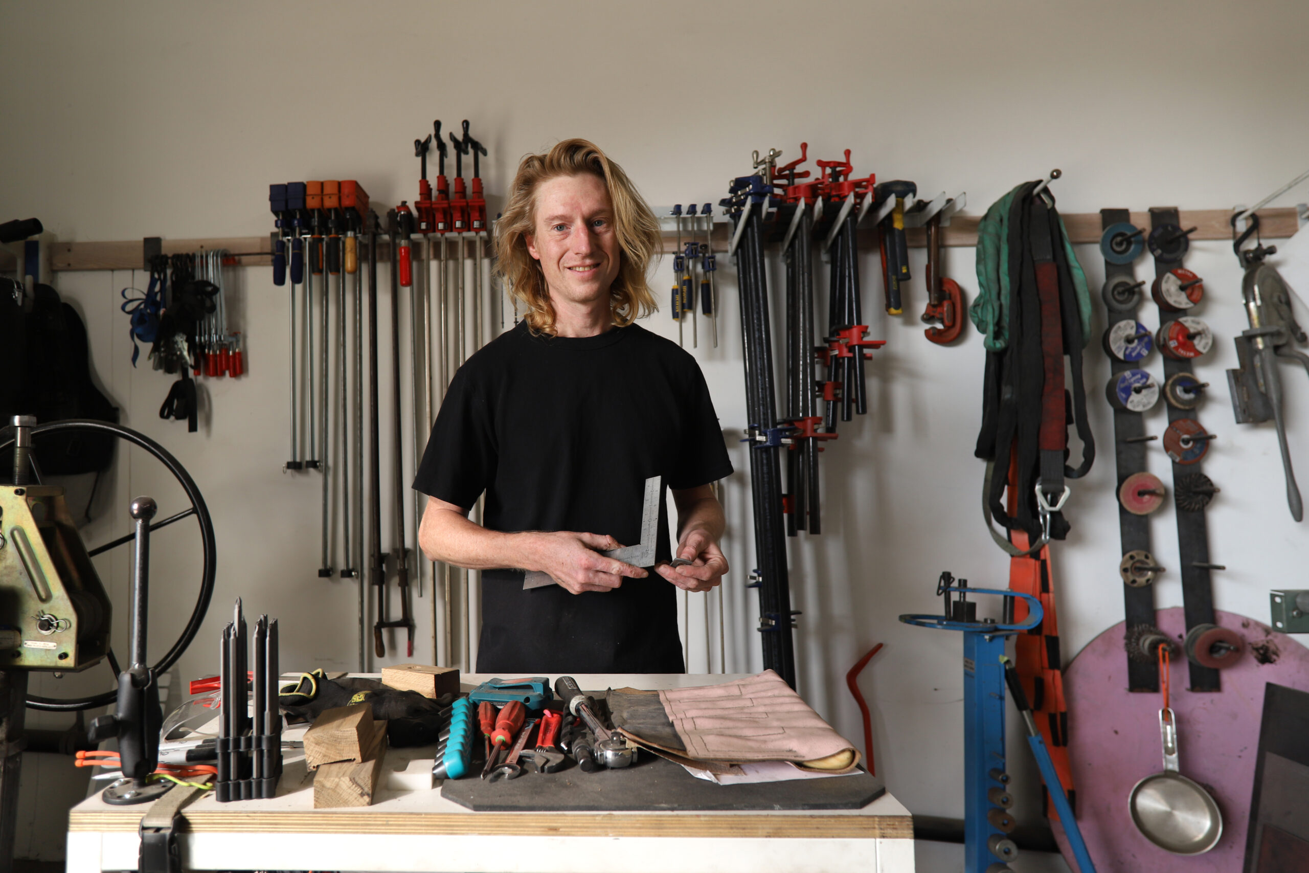 A person with long blonde hair, possibly Charles Mathis, stands smiling at a workbench in a workshop. The cluttered bench reflects their skill as a fabricator, with clamps and wrenches neatly on the wall. Perhaps they are making work for other artists here.