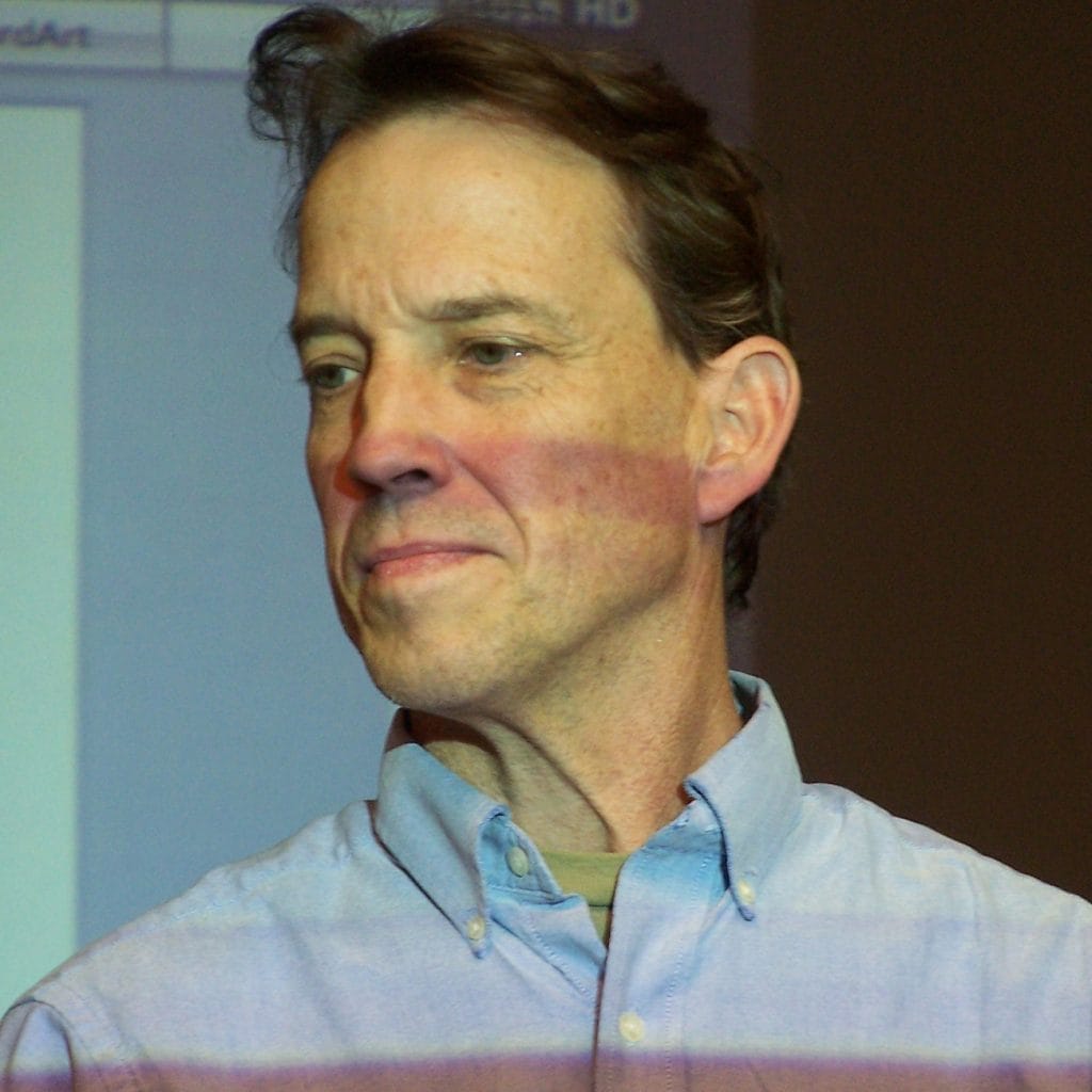 A man with short hair wearing a light blue button-up shirt is looking to the side with a neutral expression. There is a projected screen with text partially visible in the background.