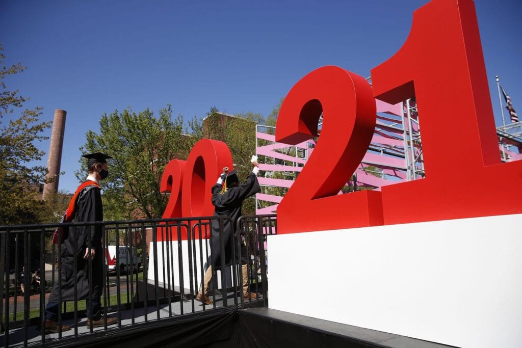 Graduates in caps and gowns walk past large red and white numbers 2021 on an outdoor stage, marking the area commencements. One graduate touches the number one. Trees and a blue sky serve as a backdrop to this memorable commencement moment.