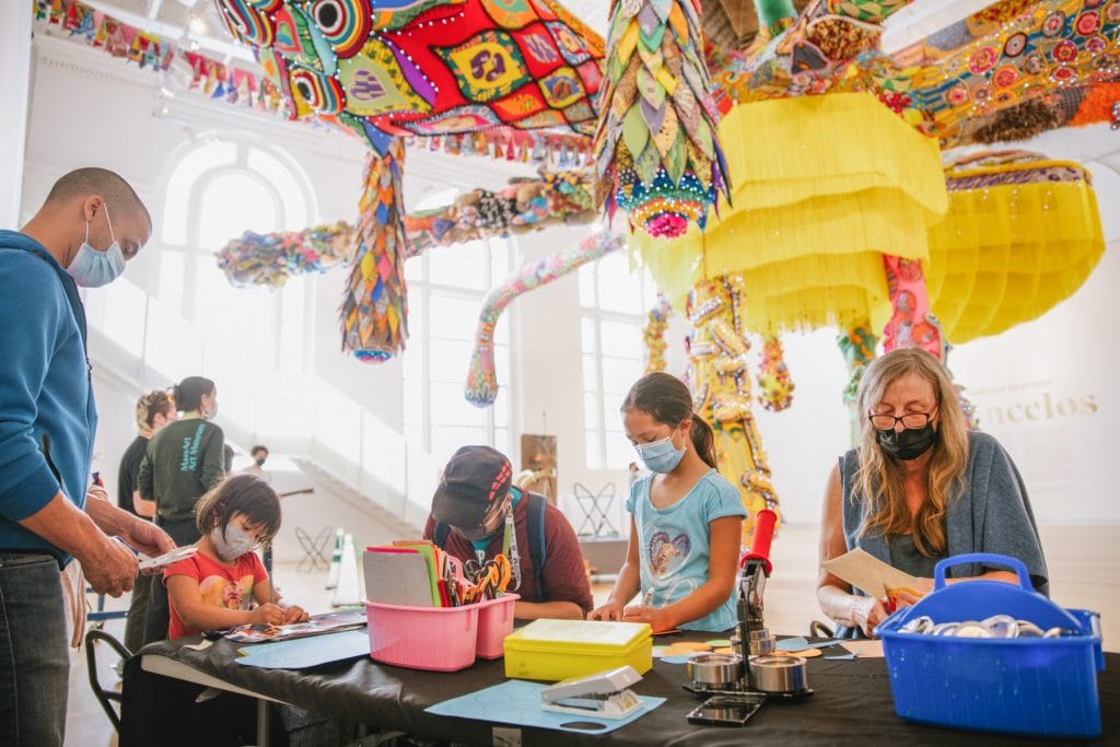 People at a table doing arts and crafts in a bright space with colorful hanging decorations. Adults and children are engaged in activities, wearing masks. Large windows and vibrant textiles create a lively atmosphere.
