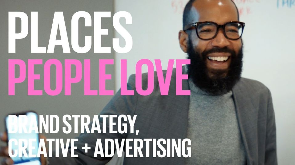 A bearded man wearing glasses and a gray jacket laughs while holding a phone. Behind him, a whiteboard showcases the message Places People Love with Brand Strategy, Creative + Advertising. His smile hints at the success of their Boston-based agency.