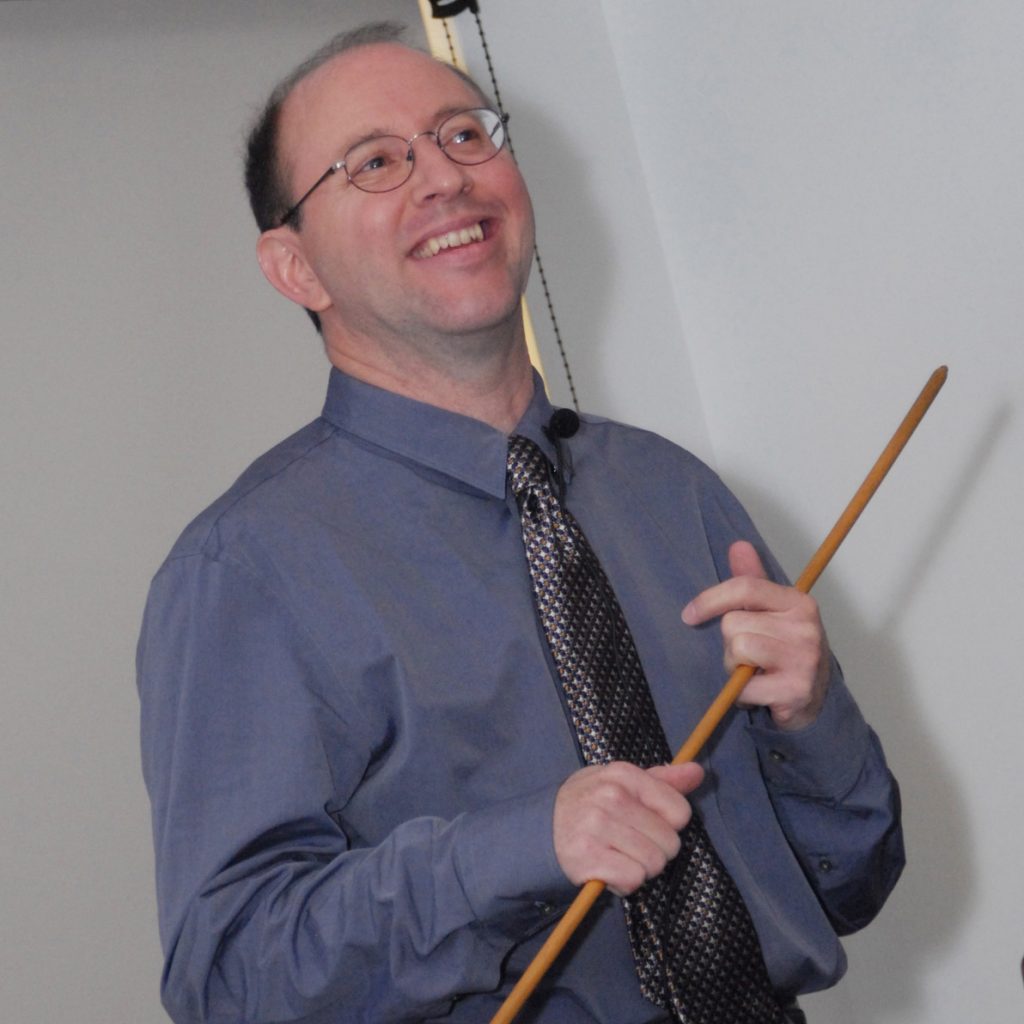 A person in a blue shirt and patterned tie is smiling while holding a wooden pointer. They appear to be engaged in a presentation or teaching session.