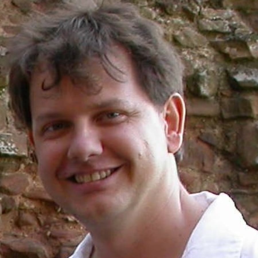 Smiling person with short, curly hair wearing a white shirt. They are standing in front of a textured stone wall.