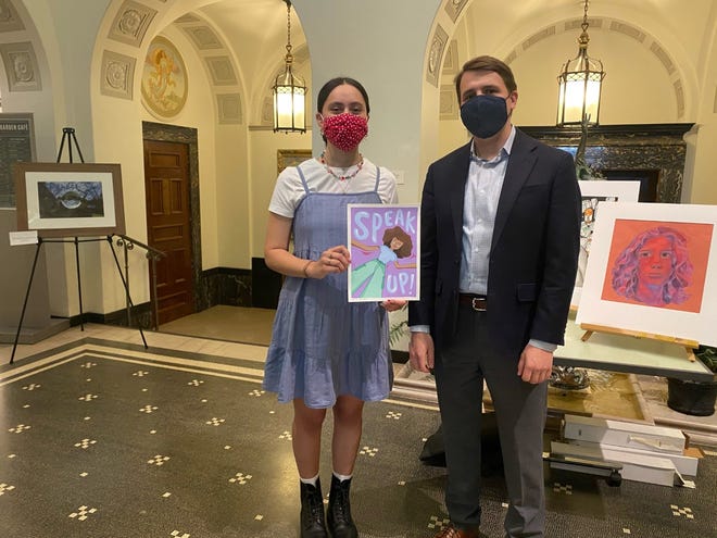 A person in a light blue dress holds a colorful Speak Up! poster next to someone in a suit at an exhibition featuring artworks from Dover High School. Both wear masks amidst the ornate architecture and patterned flooring, echoing the grandeur of the U.S. Capitol.