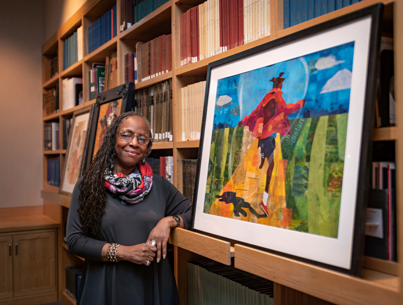 In July, a woman with long hair and glasses stands smiling beside a colorful painting in a Boston library. The artwork, part of the local arts scene, depicts a person in a red jacket with a dog against a vibrant landscape. Bookshelves fill the background.