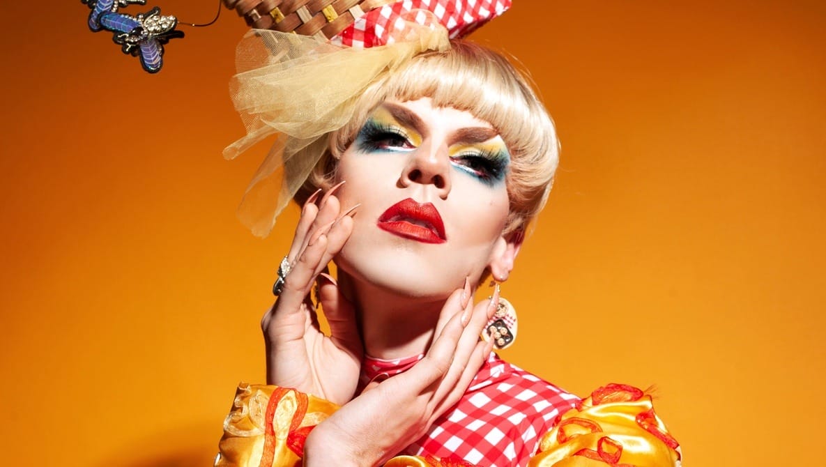 A person in vibrant drag makeup and attire strikes a pose against an orange background. Featuring bold eye makeup, red lipstick, and a blonde wig adorned with a basket, their ensemble is completed by Eric Magnussens photography capturing the red and white checkered outfit with large earrings and embellishments.