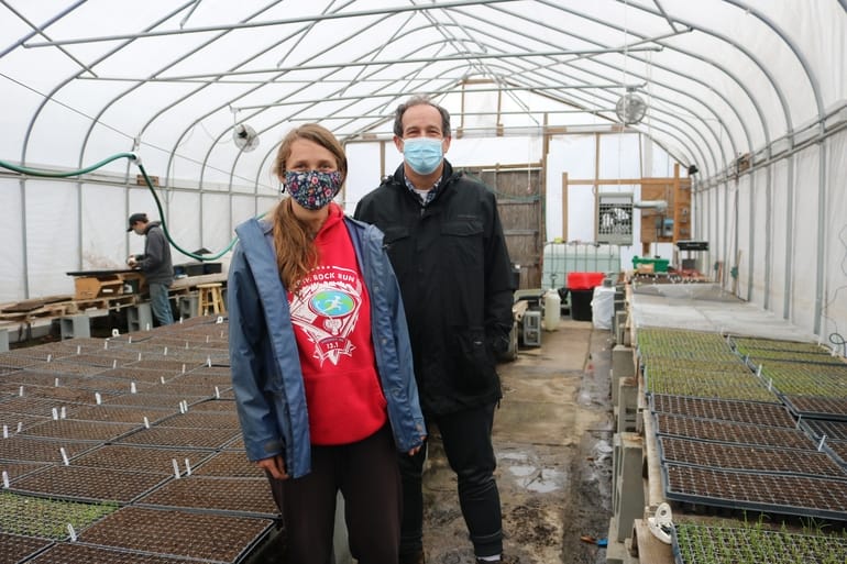 Two sustainably minded young farmers wearing face masks stand inside a greenhouse brimming with trays of seedlings. The person on the left sports a red hoodie, while the one on the right opts for a dark jacket. The spacious greenhouse, with its transparent walls and ceiling, is set in Central Mass.