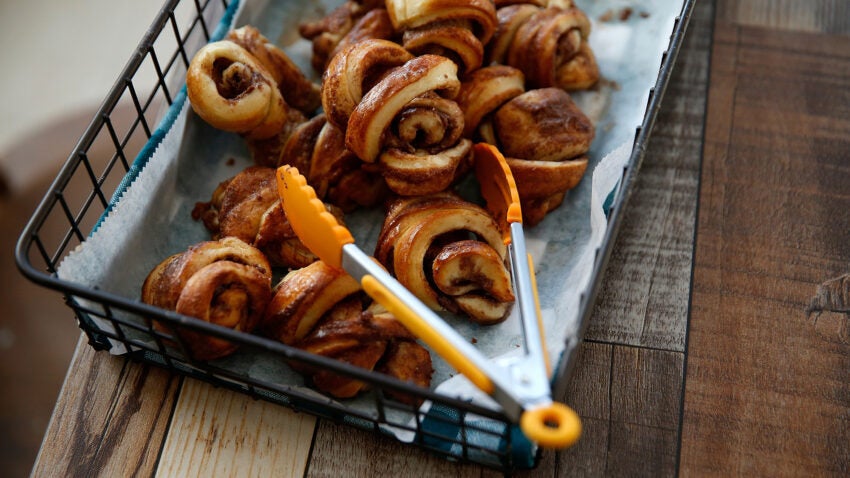 A wire basket lined with parchment paper holds several cinnamon rolls, some slightly unrolled. An orange-handled pair of tongs rests on the edge, ready to serve this delightful taste of Israel. The basket sits elegantly on a wooden surface, evoking the warmth of mezze plates.