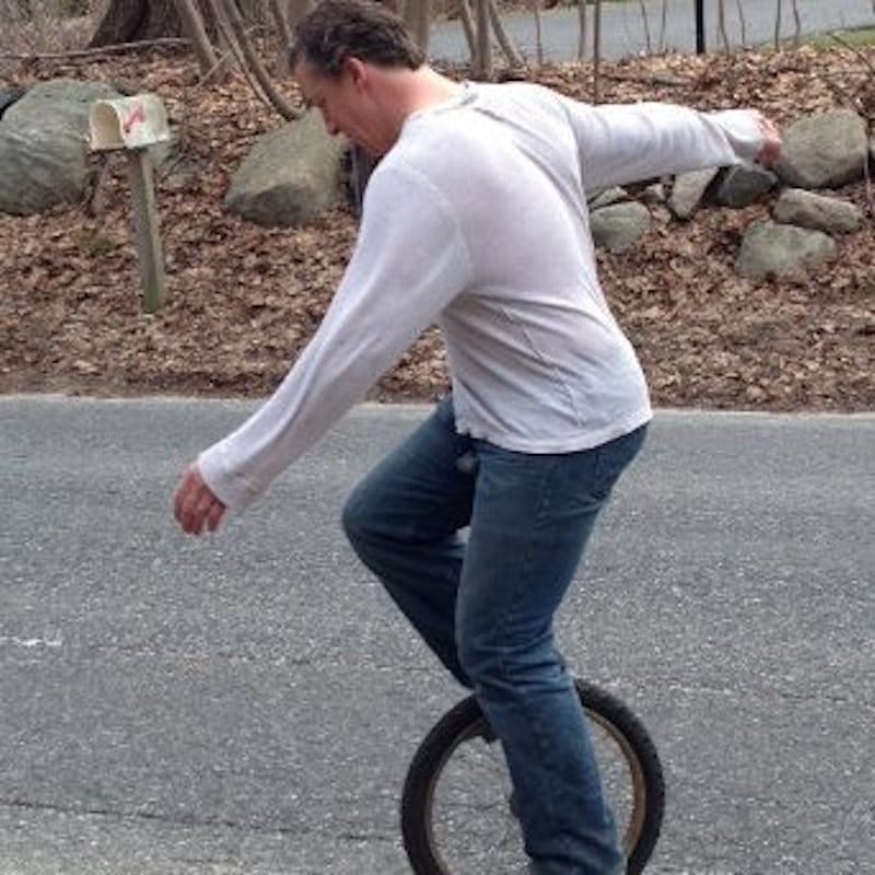 A person wearing a white long-sleeve shirt and jeans rides a unicycle on a paved road. There are rocks and fallen leaves by the roadside, and a mailbox is visible in the background.