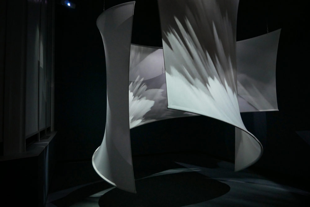 Abstract, curved fabric panels hang in a dark room, featuring projected light and shadow patterns. This captivating art installation mirrors the dynamic visual effects of a hurricane, with swirling forms symbolizing the tumultuous impact of climate change against the contrasting dark background.