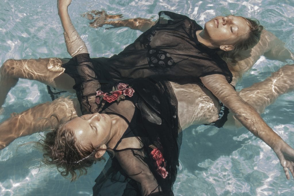 Two individuals float on their backs in a sunlit pool, wearing sheer black outfits with floral designs. The waters surface reflects light, creating patterns on their skin.