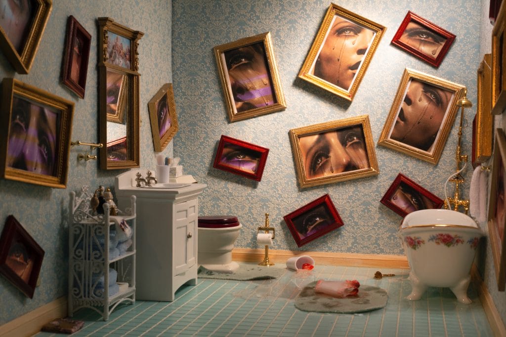 A surreal bathroom scene unfolds with pale blue patterned walls, skewed framed portraits of eyes and faces watching over a white sink, floral bathtub, and fiery red toilet. The tiled floor is a scattering of items—a houses poignant tales told through an enigmatic fallen frame.