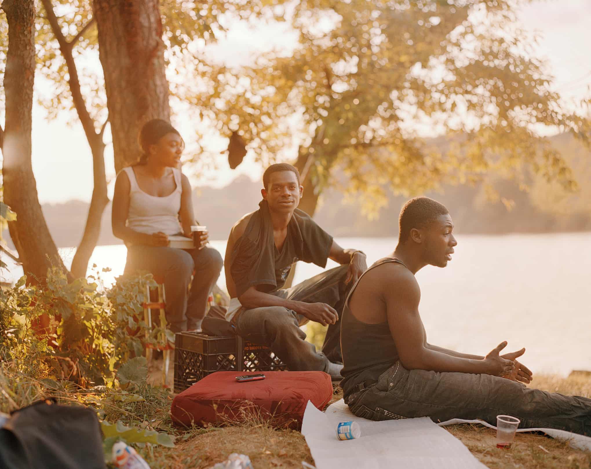Three people relax by a lakeside in warm, golden light in Irina Rozovskys Best Photograph. One sits on a crate, another reclines on the ground. Various drinking cups and a red cushion are nearby. Trees and water provide a tranquil backdrop in this World in One Park scene.