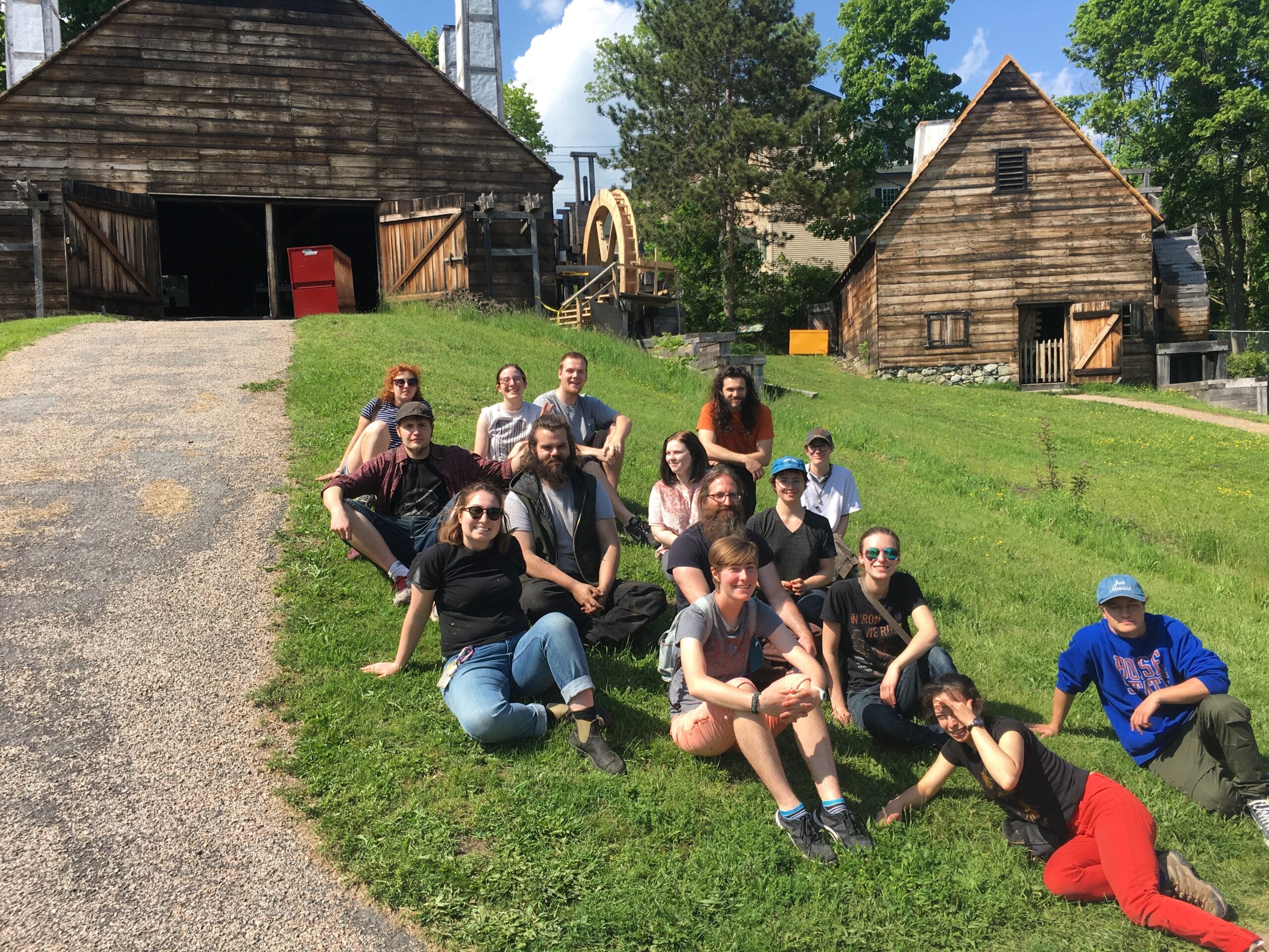 A group of MassArt students sits on a grassy hillside under a sunny sky. Behind them, rustic wooden buildings and greenery complement the scene. Relaxed and smiling in casual attire, they enjoy the rural, historic ambiance akin to Saugus Iron Works.