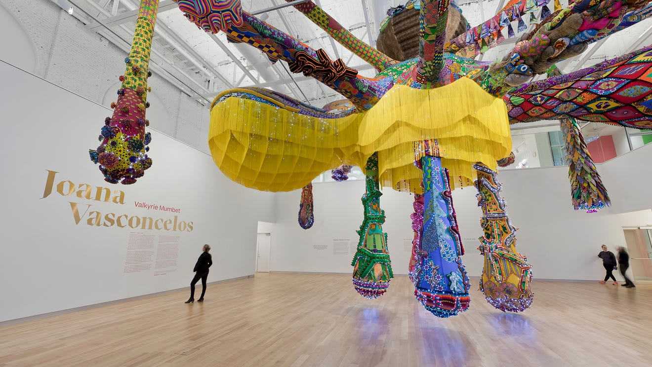 A vibrant, large-scale installation by Joana Vasconcelos is suspended from the white gallery ceiling of the MassArt Art Museum. It features colorful, intricate patterns and textures. People observe the artwork, which dominates the spacious room with its captivating presence.