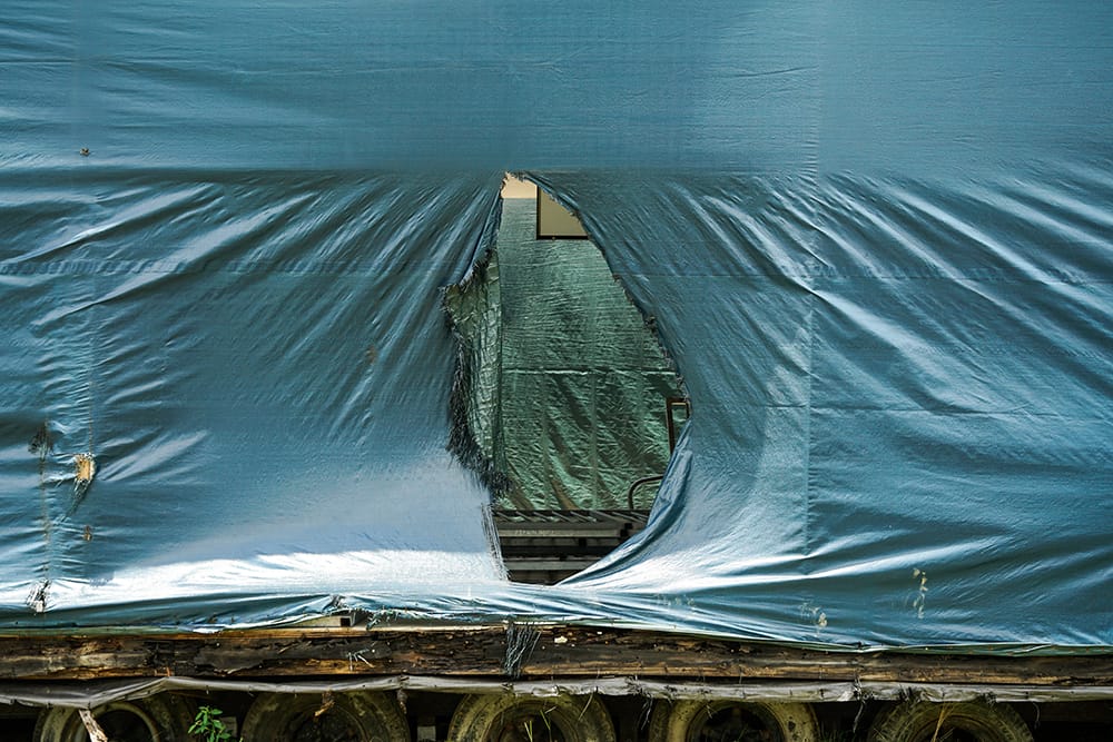 A large, blue tarp covering a structure has a jagged, circular tear, revealing another tarp inside. Below are stacked tires and wooden beams. The wear on the tarp suggests exposure to the elements, mirroring scenes from Leah Dyjaks award-winning photographic themes.