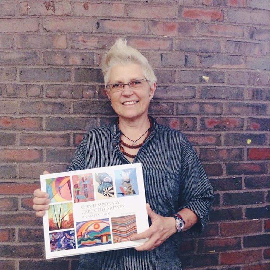 A person with short, light hair and glasses stands smiling against a brick wall. They hold a book titled Contemporary Cape Cod Artists: On Abstraction, its colorful cover exuding a sense of creative wellness and vibrancy.