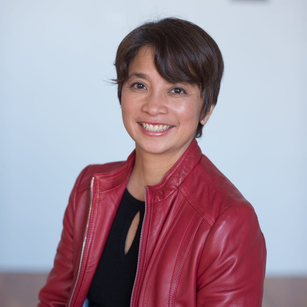 A person with short dark hair is smiling and wearing a red leather jacket over a black top. They are sitting in front of a light blue background.